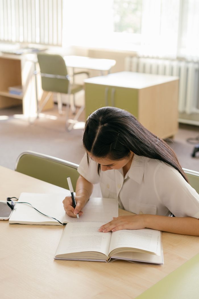 Home Young woman writing in a notebook, studying at a library table.